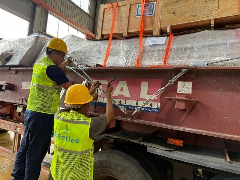 Two logistics workers in yellow safety vests tightening steel wire ropes and turnbuckles to secure wrapped industrial cargo on a flatbed truck.