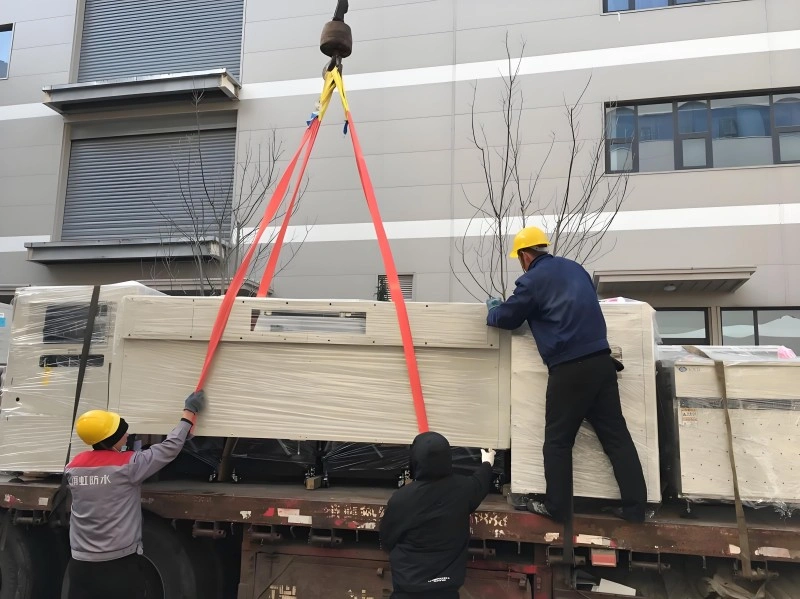 Industrial workers in yellow hard hats guiding a large machine wrapped in protective plastic during a crane lift onto a flatbed truck, demonstrating safe rigging practices.