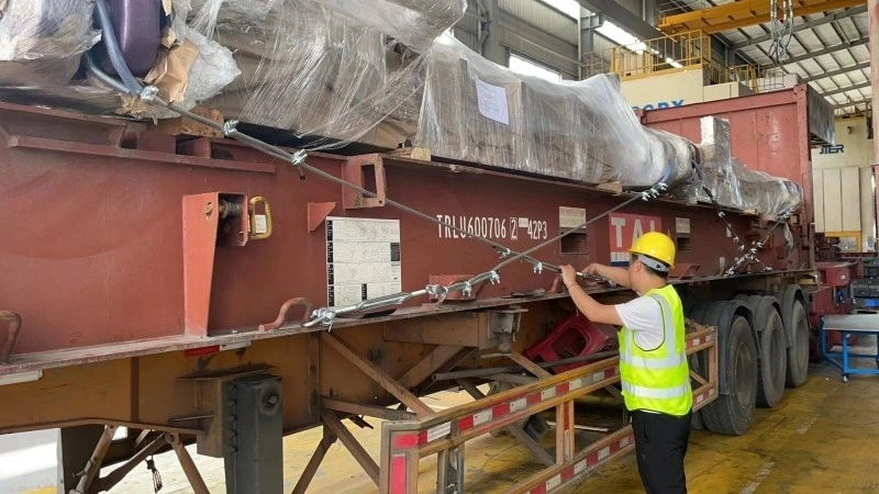 A worker in a yellow hard hat and safety vest tightening steel wire lashing on a heavy industrial machine wrapped in plastic, loaded on a flat rack container in a factory setting.