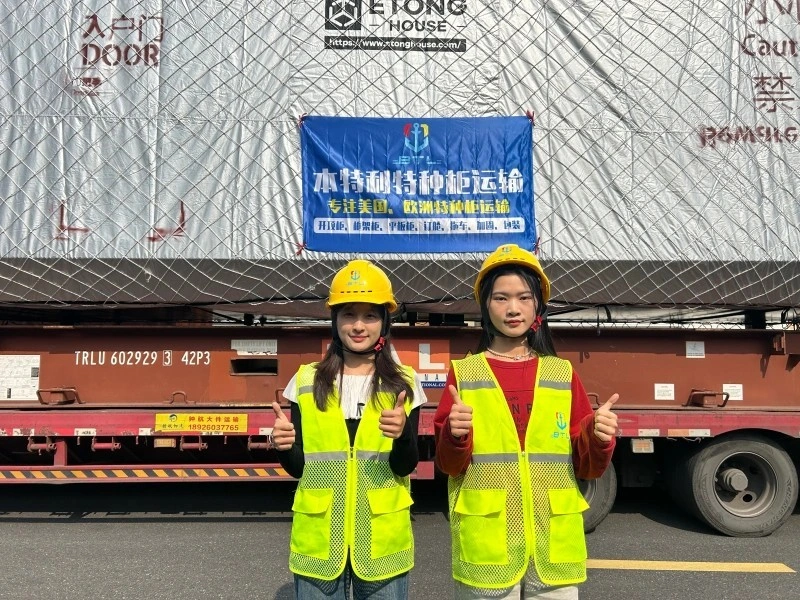 Two female logistics coordinators in yellow safety vests and hard hats giving thumbs up in front of a large cargo truck with a banner advertising specialized transportation services to the US and Europe.
