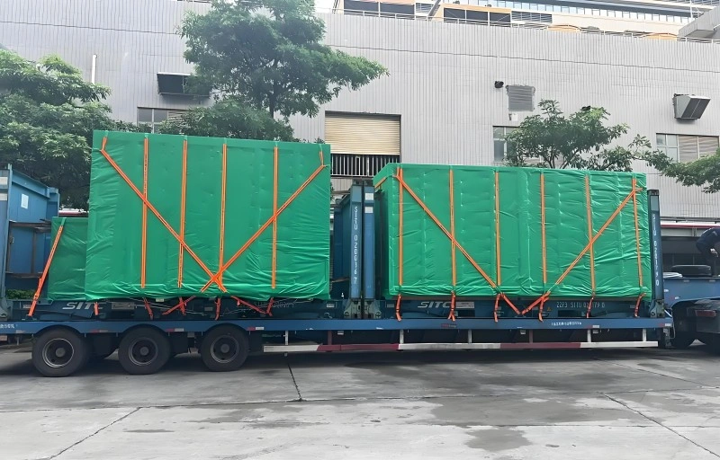 A logistics worker standing on a flat rack trailer securing a large green waterproof tarp over oversized industrial machinery, demonstrating proper sea freight packaging and lashing procedures for heavy equipment transport.
