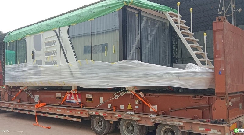 A large prefabricated glass module wrapped in white stretch film and green tarp, secured with orange lashing straps on a flatbed trailer at a logistics yard.