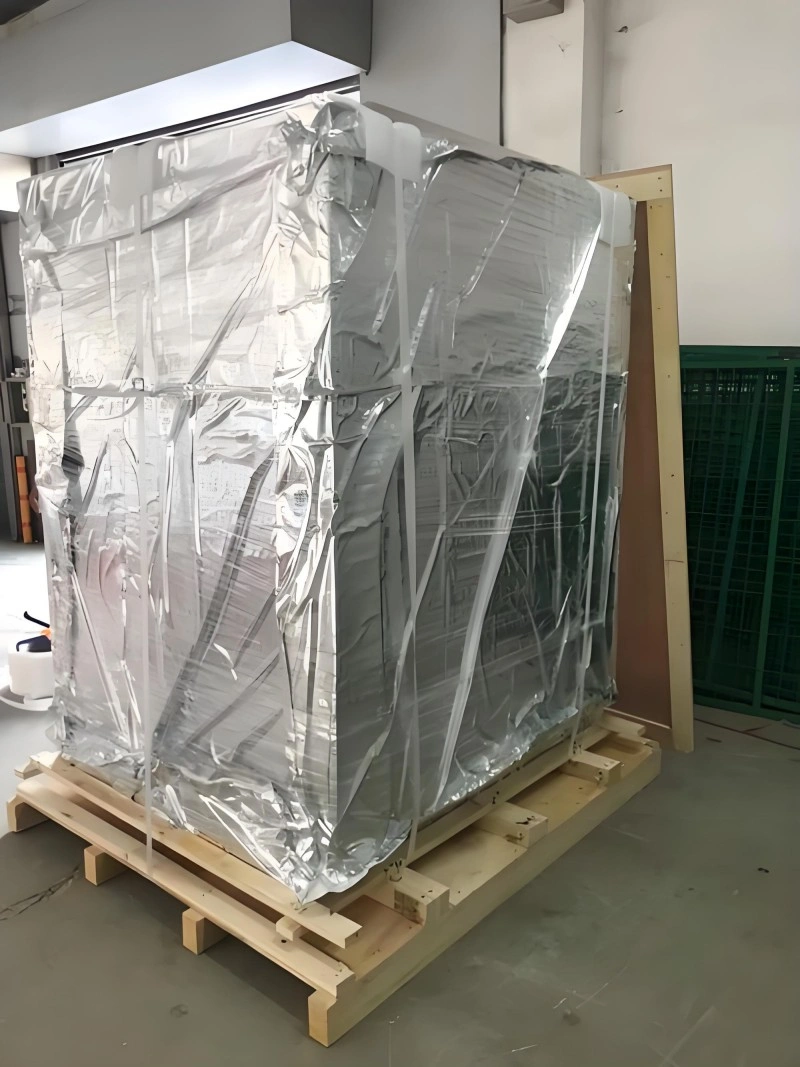 A tall rectangular machine wrapped in silver aluminum foil vacuum film and secured on a wooden pallet inside a warehouse, prepared for international shipment with full moisture and dust protection.