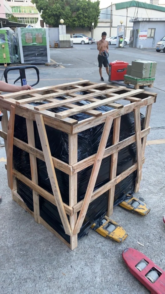 A heavy-duty wooden crate with diagonal bracing and black waterproof wrapping being moved by a pallet jack, showcasing robust protection for sensitive cargo.