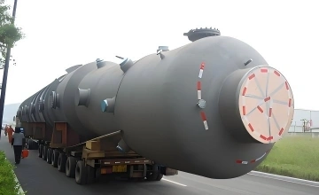 Oversized pressure vessel moving on a specialized trailer along a highway, with a city skyline in the background, representing the journey from factory to port
