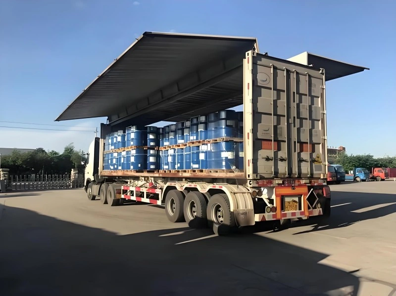 Blue chemical drums loaded on wooden pallets inside an open-top shipping container mounted on a flatbed truck
