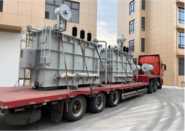 Red low-bed trailer carrying two large grey power transformers secured with heavy-duty straps for safe OOG transportation