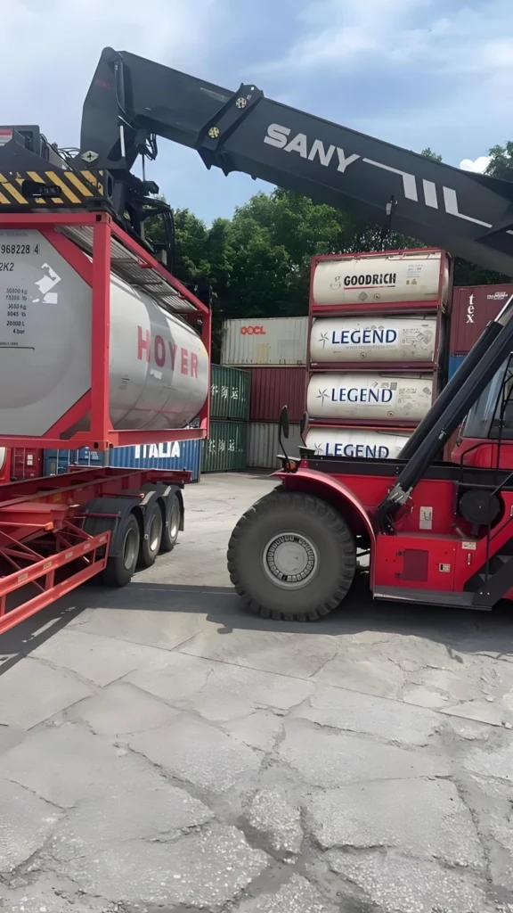A red SANY reach stacker is lifting a red-framed ISO tank container with "HOYER" branding onto a flatbed trailer. In the background, multiple stacked ISO tank containers from "GOODRICH" and "LEGEND" are visible in a container yard under a partly cloudy sky.
