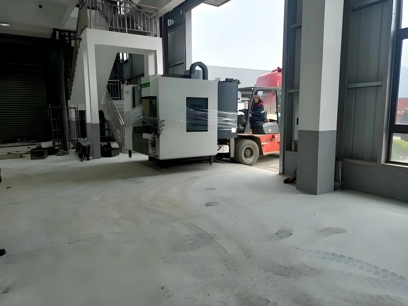 A large industrial machine wrapped in protective plastic film being carefully maneuvered by a red forklift through a factory doorway during a plant move.