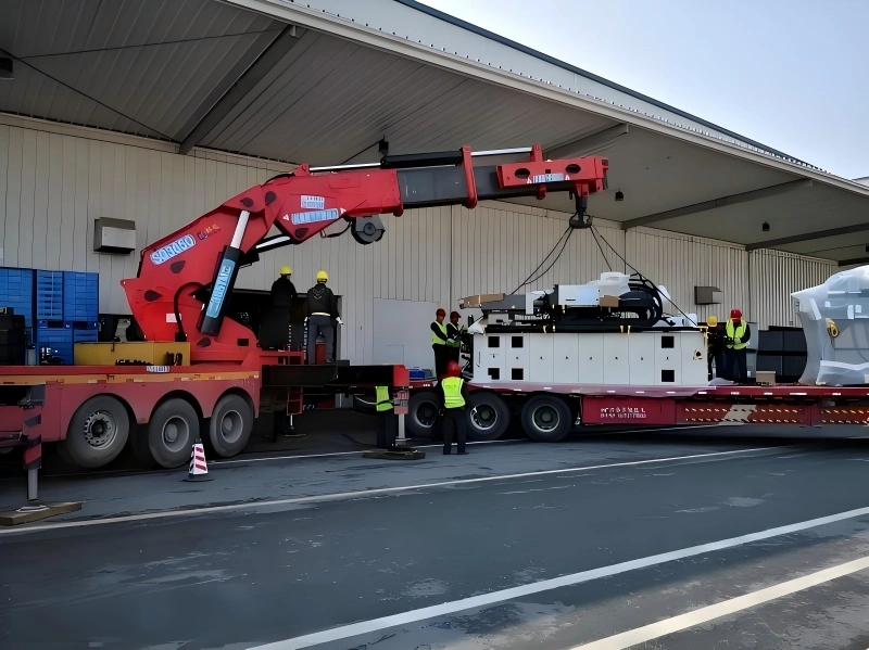 A red mobile crane carefully lifting a large white industrial unit onto a flatbed truck, with workers in high-visibility vests guiding the operation at an outdoor loading area.