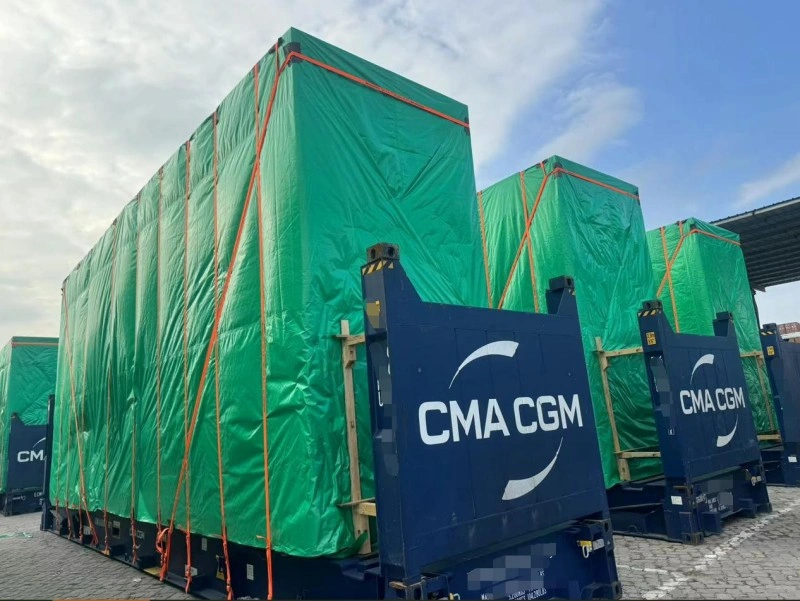 Large industrial machinery wrapped in green protective tarpaulin and secured with orange lashing straps on CMA CGM flat rack containers at a port terminal.