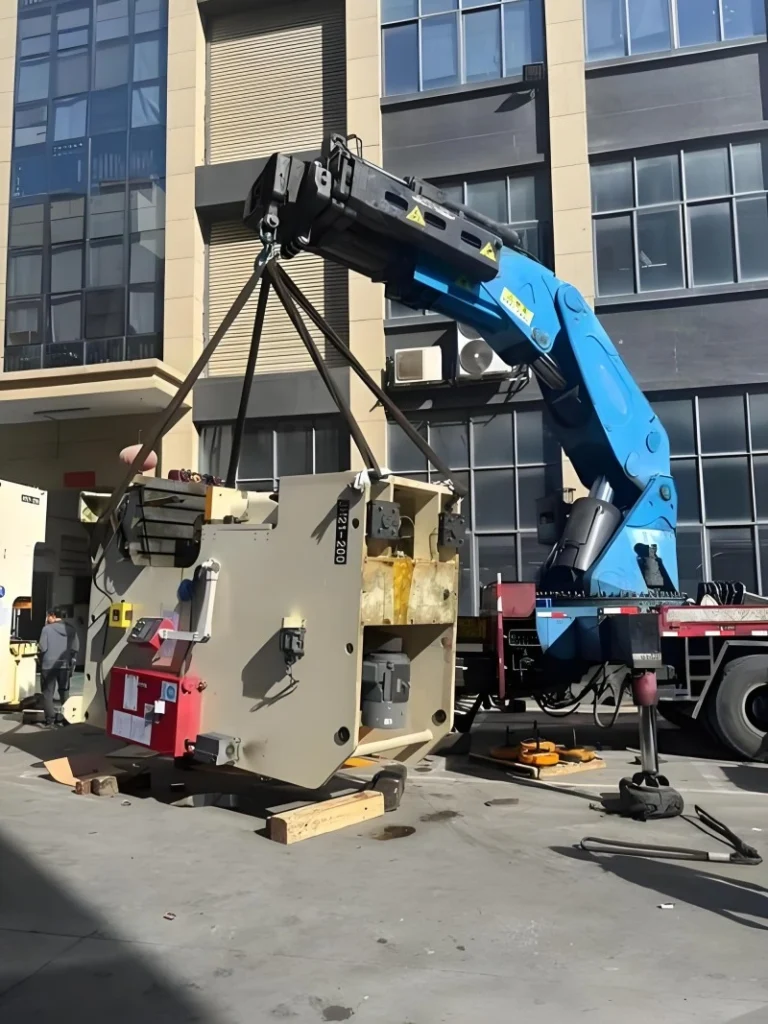 A massive blue and white CNC machine being carefully positioned onto a multi-axle flatbed trailer using overhead lifting gear in an industrial facility.