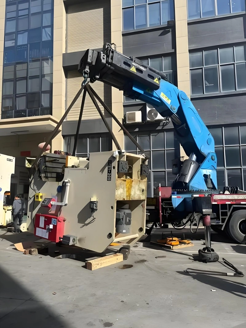 A massive blue and white CNC machine being carefully positioned onto a multi-axle flatbed trailer using overhead lifting gear in an industrial facility.