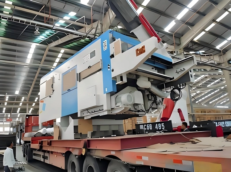 A massive blue and white CNC machine being carefully positioned onto a multi-axle flatbed trailer using overhead lifting gear in an industrial facility.