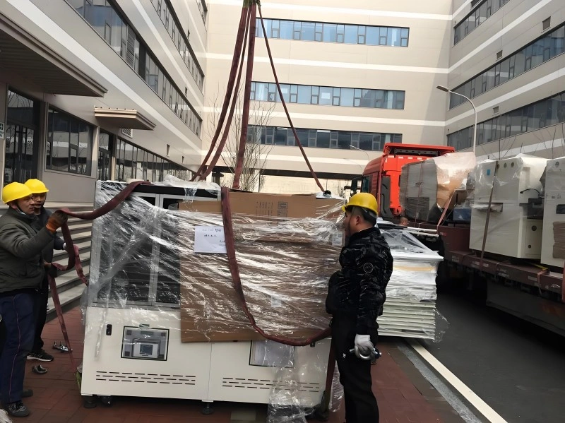 A large white precision machine wrapped in protective plastic film being carefully maneuvered by workers with lifting straps near a transport truck.