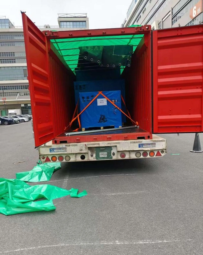 The rear view of an open shipping container showing a blue industrial machine securely strapped with orange straps and covered with green tarpaulin.