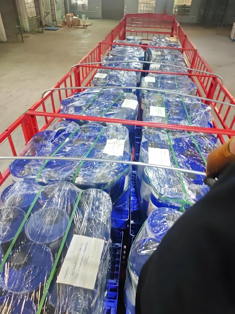 Blue plastic chemical containers wrapped in clear plastic film and secured with green straps on blue pallets inside a red truck bed