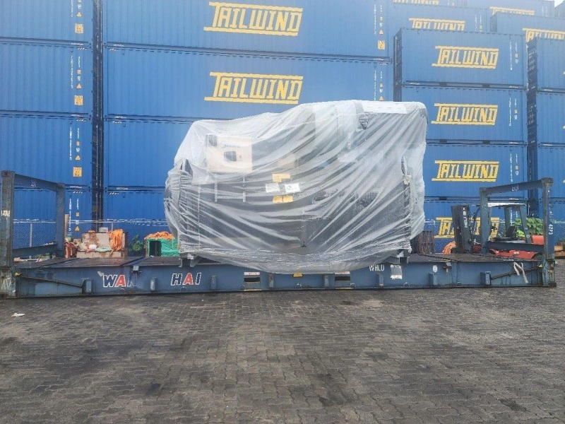 A large industrial machine wrapped in clear plastic is loaded onto a blue flat rack container in a busy shipping yard, with multiple blue shipping containers stacked in the background. The cargo is properly secured with lashing equipment.