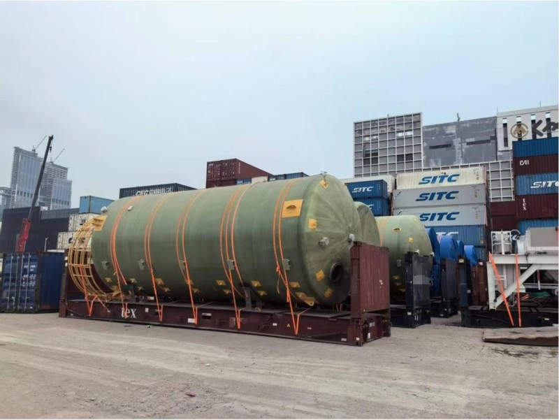 A large green fiberglass tank strapped securely onto a flat rack container, surrounded by stacked shipping containers, demonstrating safe lashing techniques for OOG cargo during plant moves