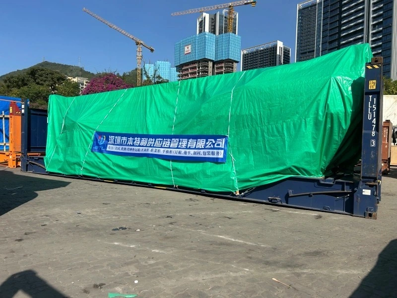 A large, tarp-covered cargo on a flat rack container with a banner for Shenzhen BTL Supply Chain Management Co., Ltd., showcasing their OOG shipping services in an urban port area.