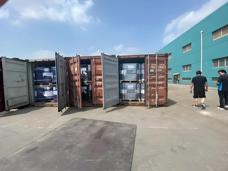Three open shipping containers on an outdoor concrete pad, loaded with pallets of blue and black industrial drums. Two workers in dark clothing are walking near the containers, with a large teal warehouse building in the background under a partly