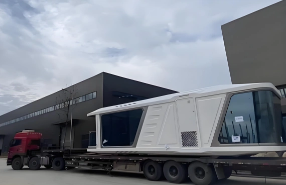 Side perspective of a space capsule cabin on a flatbed trailer outside a factory, ready for inland transport to the port