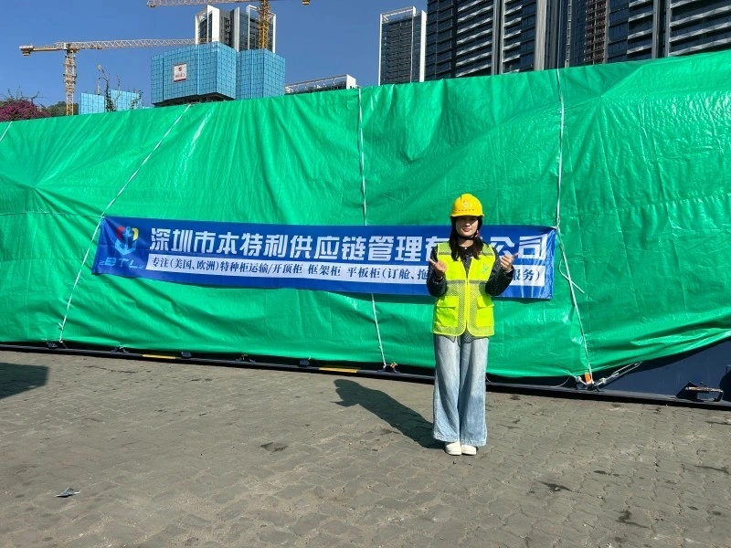 A female logistics professional in a yellow hard hat and safety vest stands smiling and giving a thumbs-up in front of a massive cargo load covered with a green tarpaulin. A blue banner with Chinese text identifying "Shenzhen BTL Supply Chain Management Co., Ltd." is visible on the cargo. In the background, there are high-rise buildings and a construction crane