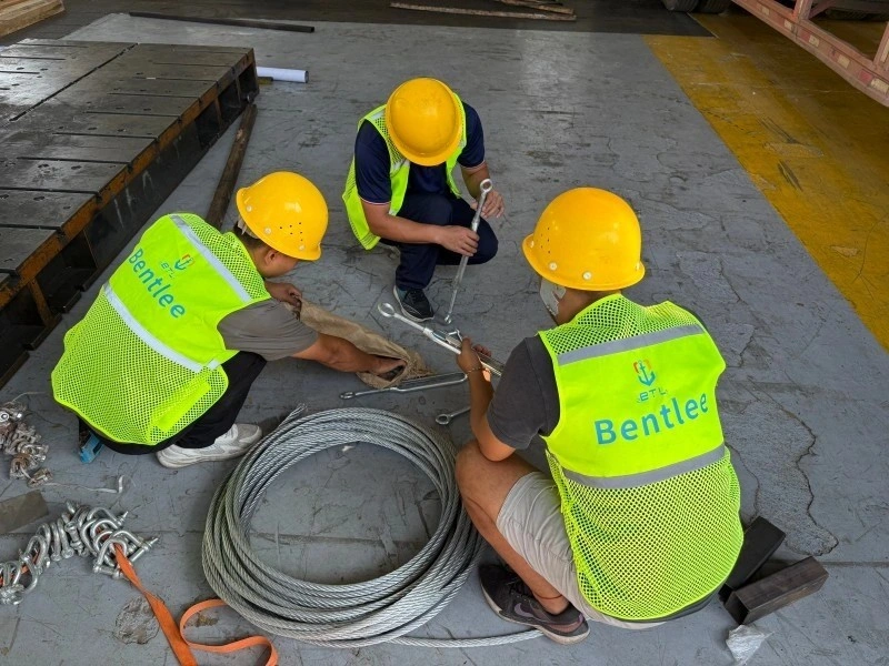 Three workers in yellow hard hats and Bentlee safety vests inspecting and organizing steel wire ropes, shackles, and turnbuckles on a warehouse floor