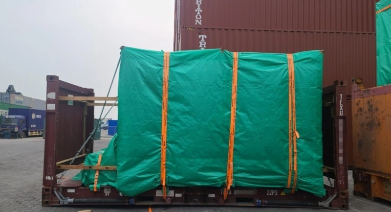 A large, rectangular cargo unit is covered with a green waterproof tarp and secured on a brown flat rack container using multiple orange lashing straps, with wooden support frames visible on the sides, set against a backdrop of stacked shipping containers in a port yard.