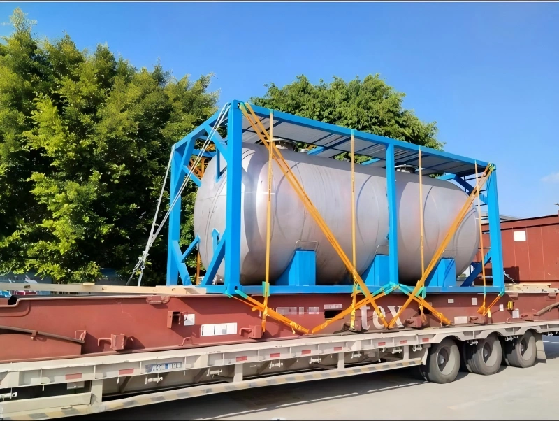 A side view of a large industrial tank, housed in a blue steel frame, being transported on a red flatbed trailer. The cargo is secured with crisscrossing orange straps. The background shows green trees and a clear blue sky, indicating good weather conditions for transport.
