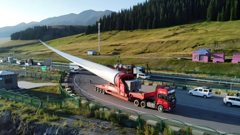 A red multi-axle lowbed trailer carries an 84-meter-long white wind turbine blade along a winding mountain highway, with green hills and forested slopes in the background, illustrating long-distance oversized cargo transport for wind energy infrastructure.