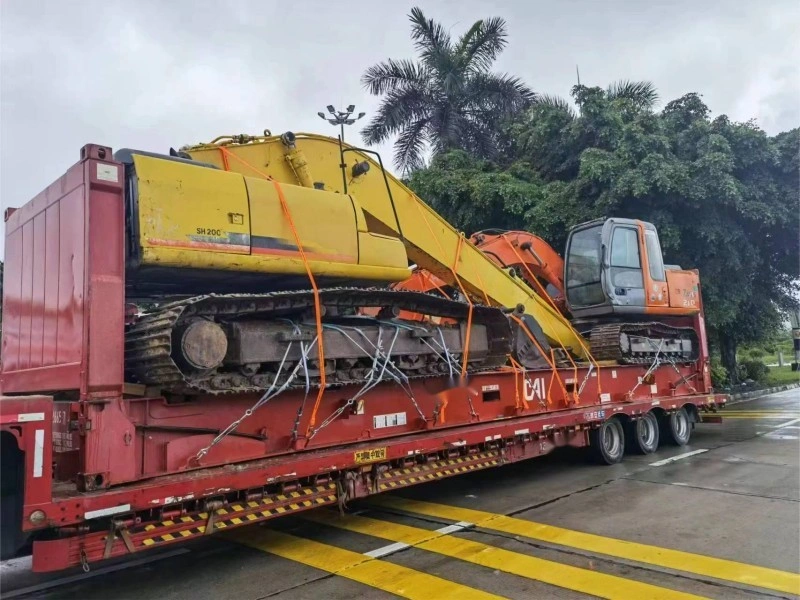 A yellow and orange tracked crane is loaded onto a red lowboy trailer for transportation. The heavy construction vehicle is secured with multiple orange straps and chains. The truck is on a wet road, with lush green trees in the background, suggesting recent rain.