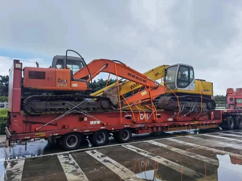 Two large excavators, one orange and one yellow, are loaded onto a red, multi-axle lowboy trailer. The heavy construction equipment is interlocked and secured with a complex web of orange straps and metal chains. The ground is wet, reflecting the vehicles, indicating it has recently rained.