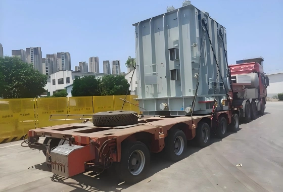 Brown extendable trailer transporting a vertically oriented grey power transformer through an urban area, requiring careful route planning