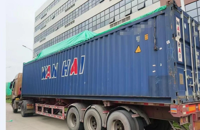 A blue Wan Hai 40ft high cube container mounted on a multi-axle flatbed truck, covered with green tarpaulin, prepared for long-distance road transport to port.