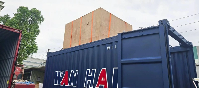 A large wooden crate secured with orange straps being loaded into a blue Wan Hai shipping container, illustrating automated and safe handling of oversized project cargo.