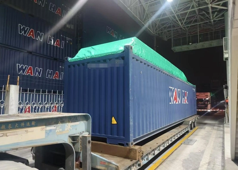 A blue Wan Hai open top container, topped with a bright green tarpaulin to shield its oversized cargo, is positioned on a low-bed trailer inside a well-lit warehouse or terminal at night, preparing for departure.