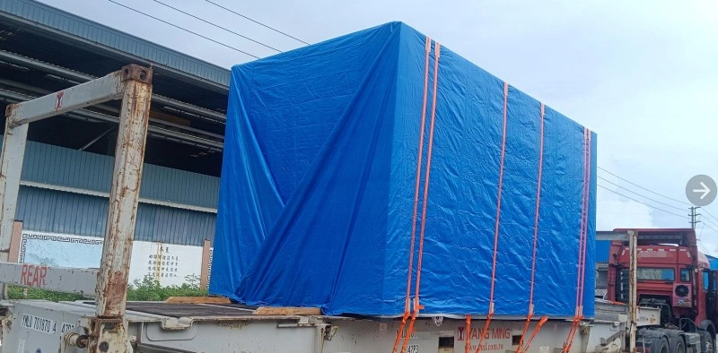 A large industrial machine wrapped in a blue waterproof tarp and secured with orange straps on a flatbed truck, ready for transport.