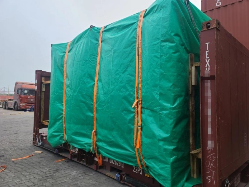 A large piece of equipment covered with a green waterproof tarpaulin and secured with orange straps on a flat rack container in a port yard.