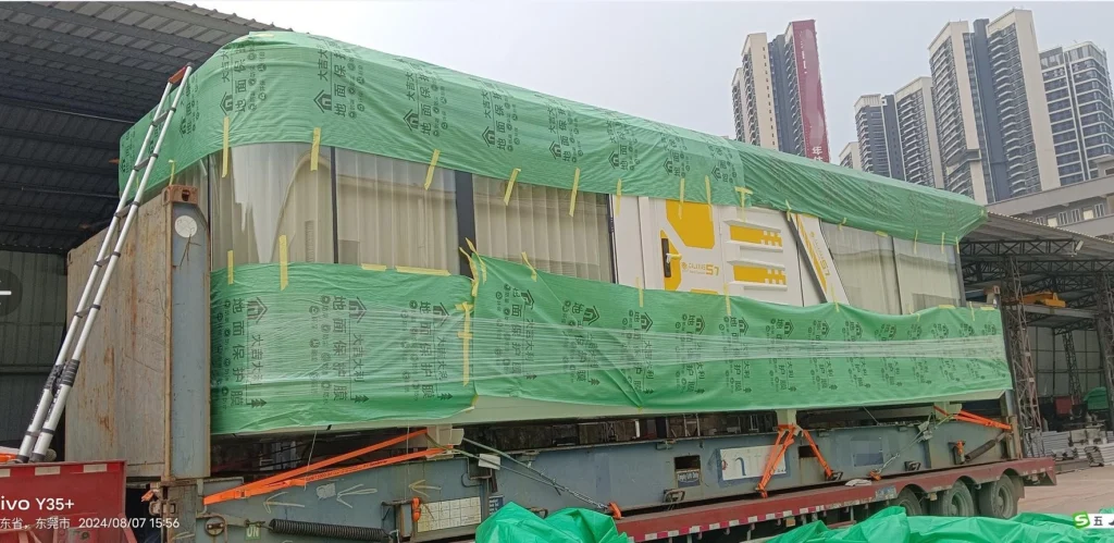 Side view of a fully wrapped oversized prefab unit covered in green protective tarpaulin, staged near urban buildings, demonstrating comprehensive weather protection for sensitive equipment.