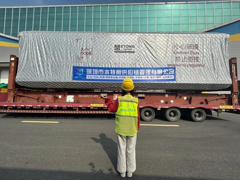 A logistics worker in a yellow hard hat and safety vest stands before a large, silver-wrapped prefabricated capsule house loaded onto a flatbed trailer, with a banner for Shenzhen Bentlee Supply Chain Management Co., Ltd. visible