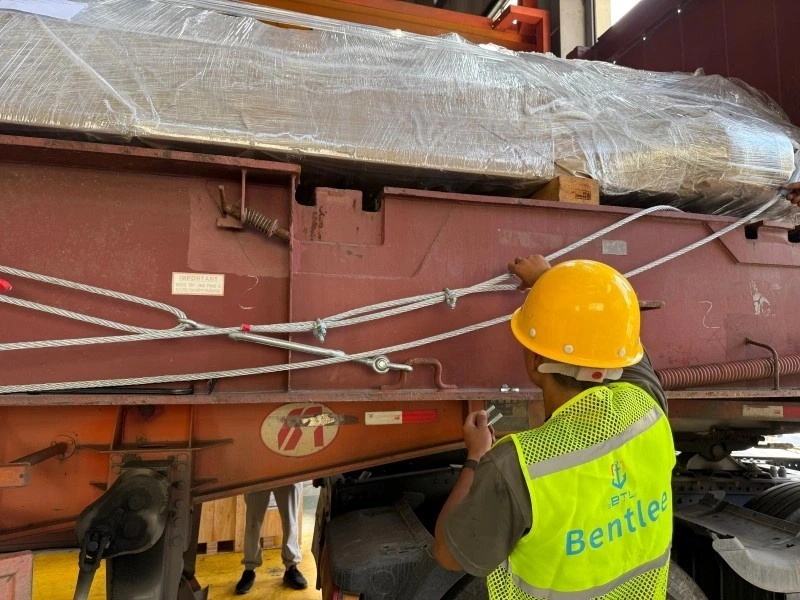 Worker in yellow hard hat and Bentlee safety vest tightening steel wire ropes to secure a large, plastic-wrapped industrial machine on a red flatbed trailer