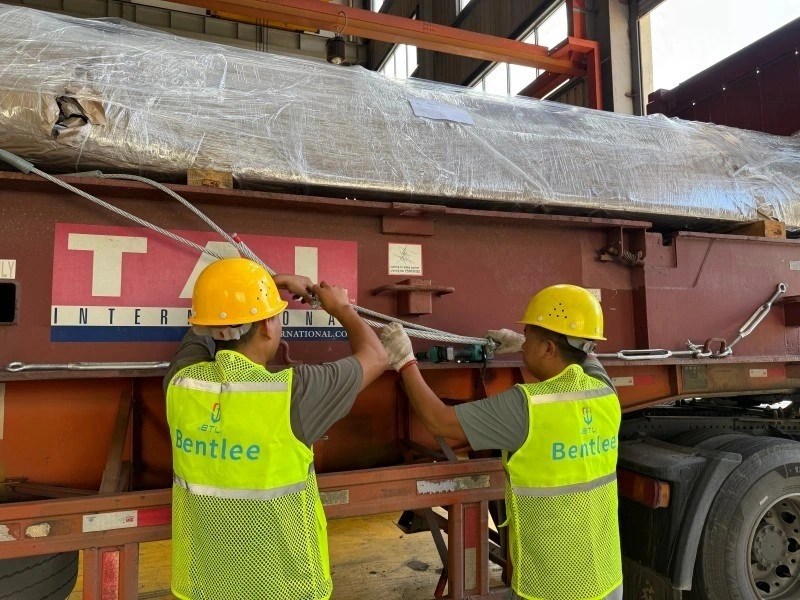 Two logistics workers wearing yellow hard hats and high-visibility vests with the "Bentlee" logo are tightening steel wire ropes to secure a long, silver-wrapped cargo item onto a large red transport frame. The operation is taking place inside a warehouse or factory setting with an overhead crane visible.
