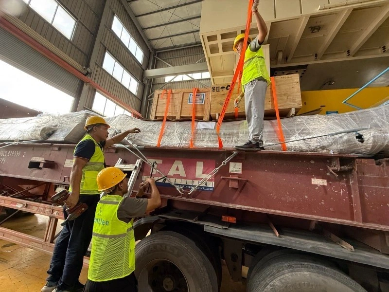 Three workers in hard hats and safety vests are securing a large, plastic-wrapped piece of machinery onto a red flatbed trailer. They are using heavy-duty orange ratchet straps and metal chains to ensure the cargo is stable for transport. The scene is inside a large warehouse or loading bay.