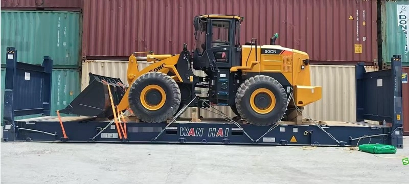 A yellow ZL50CN wheel loader is tightly strapped onto a blue flat rack container with orange lashing straps, positioned between standard shipping containers in an industrial yard, showcasing secure transport methods for heavy construction equipment.
