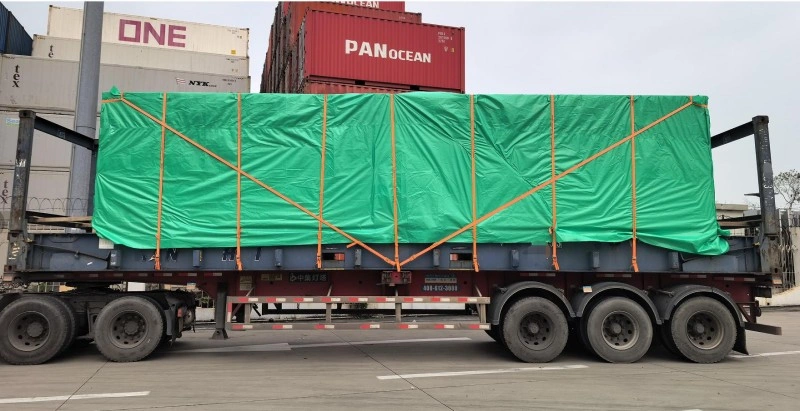 Side view of a large open top container fully covered with green tarp and secured with yellow straps, mounted on a multi-axle flatbed trailer in a port setting