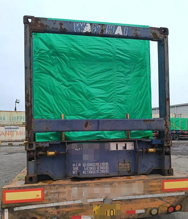 Rear view of a blue Wan Hai open top container with green tarp cover, mounted on a heavy-duty flatbed trailer, showing weight specifications and rusted frame