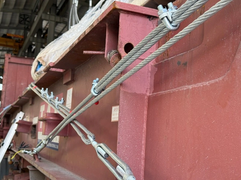 Close-up of galvanized wire rope and turnbuckles securing a large red steel structure, demonstrating professional lashing techniques for heavy automotive mold transport.