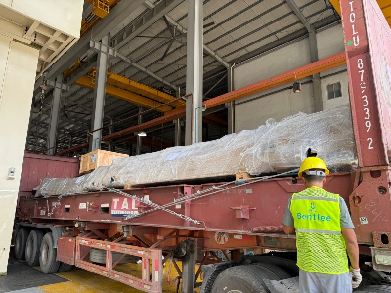 A logistics supervisor in a yellow Bentlee vest overseeing the loading of a massive shrink-wrapped automotive stamping die onto a TAL flat rack container inside a warehouse.