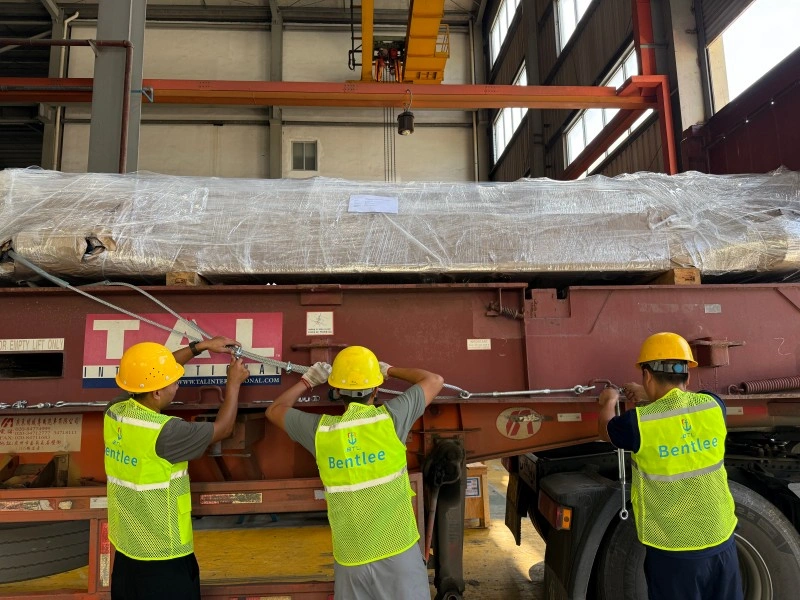 Three workers in safety helmets and Bentlee vests tightening steel cables on a shipping container, illustrating the manual labor involved in securing OOG cargo for international transport.